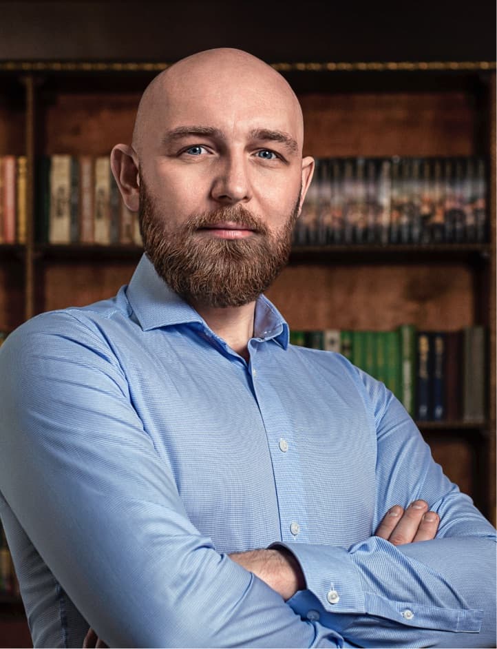 A man with a bald head and beard poses confidently in front of a bookshelf filled with books